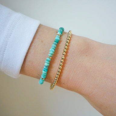 Birthstone Bead Bracelet In Turquoise next to gold ball stretch bracelet styled on wrist of model with white sleeve